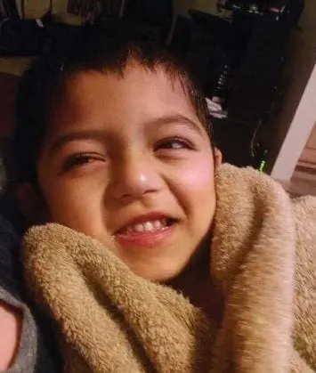 A young boy with dark hair smiles, wrapped in a tan towel.
