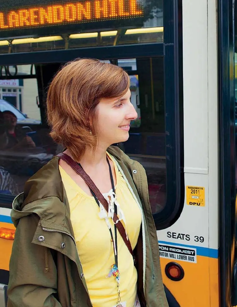 A young woman wearing a yellow shirt and olive green jacket stands in front of a bus.