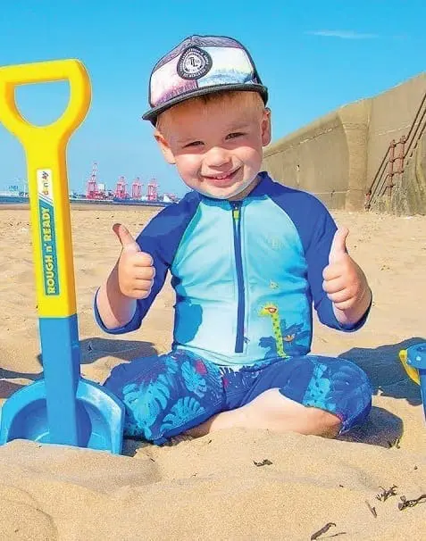 Young boy wearing a blue rash guard and swim trunks gives two thumbs up on a sandy beach next to a toy shovel.