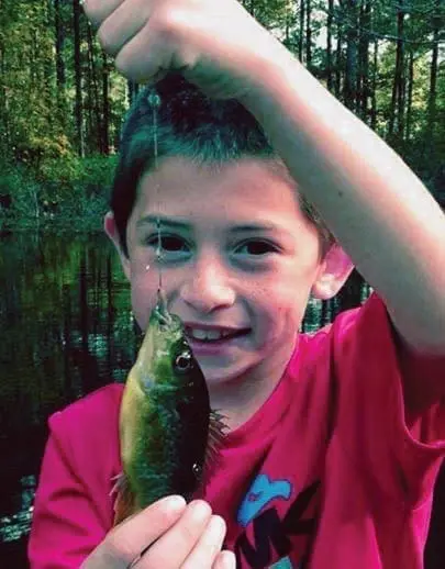 A young boy in a pink shirt holds up a small fish he caught.