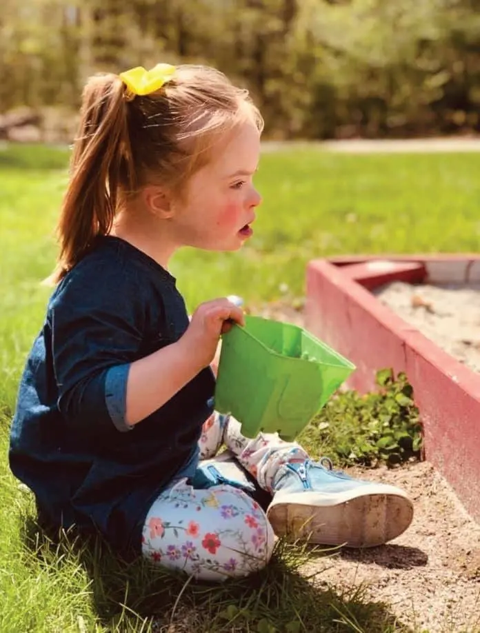 A young girl with Down syndrome sits in the grass holding a green bucket.
