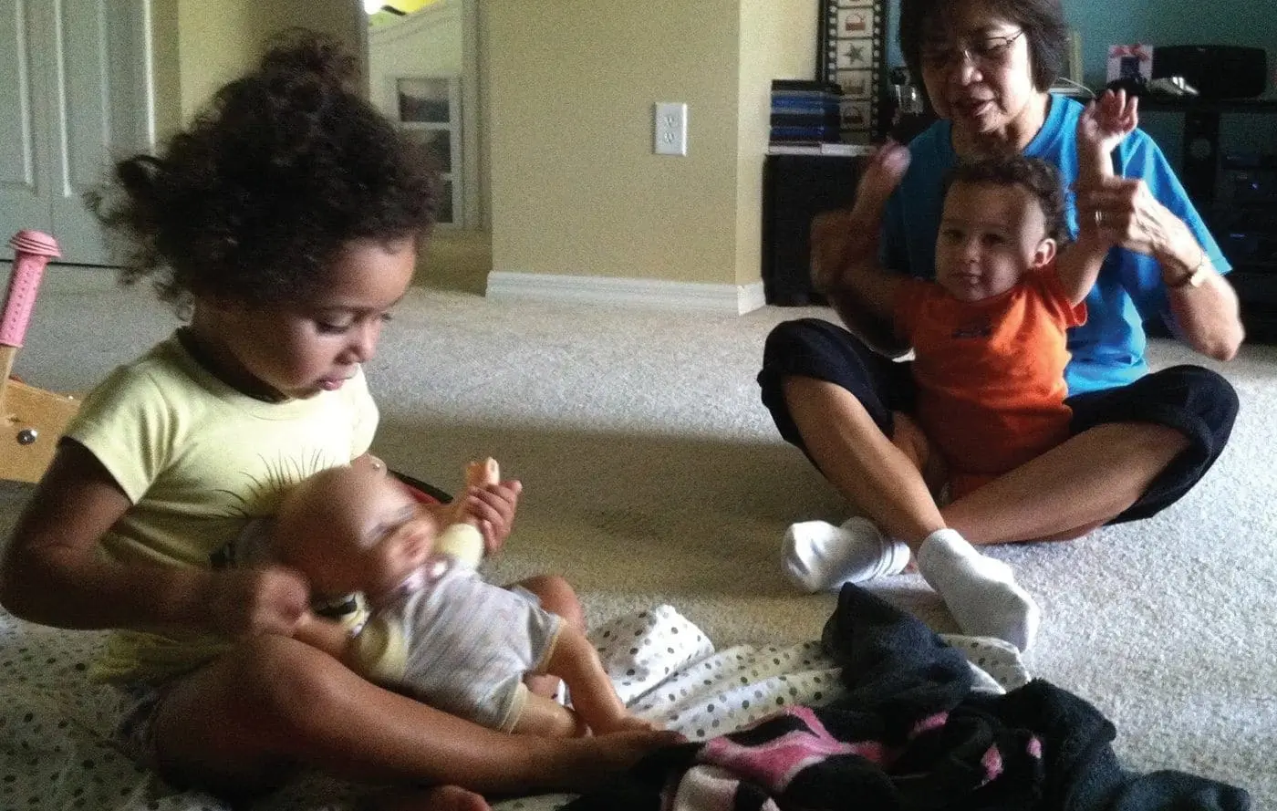 A young girl plays with a baby doll while a woman and another baby sit on the floor behind them.