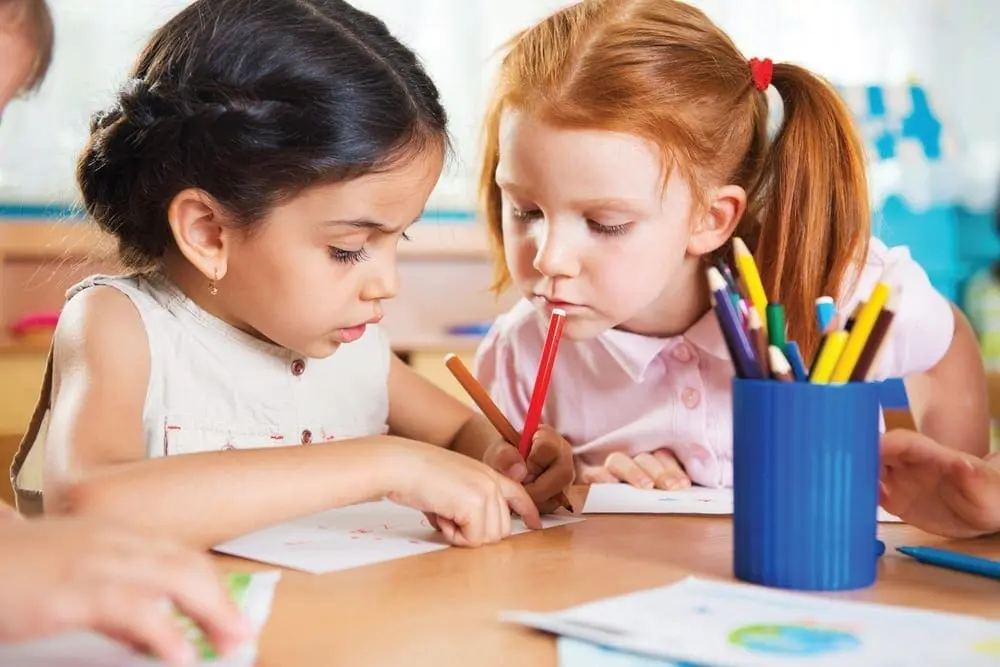 Two young girls are drawing and coloring at a table with a teacher.