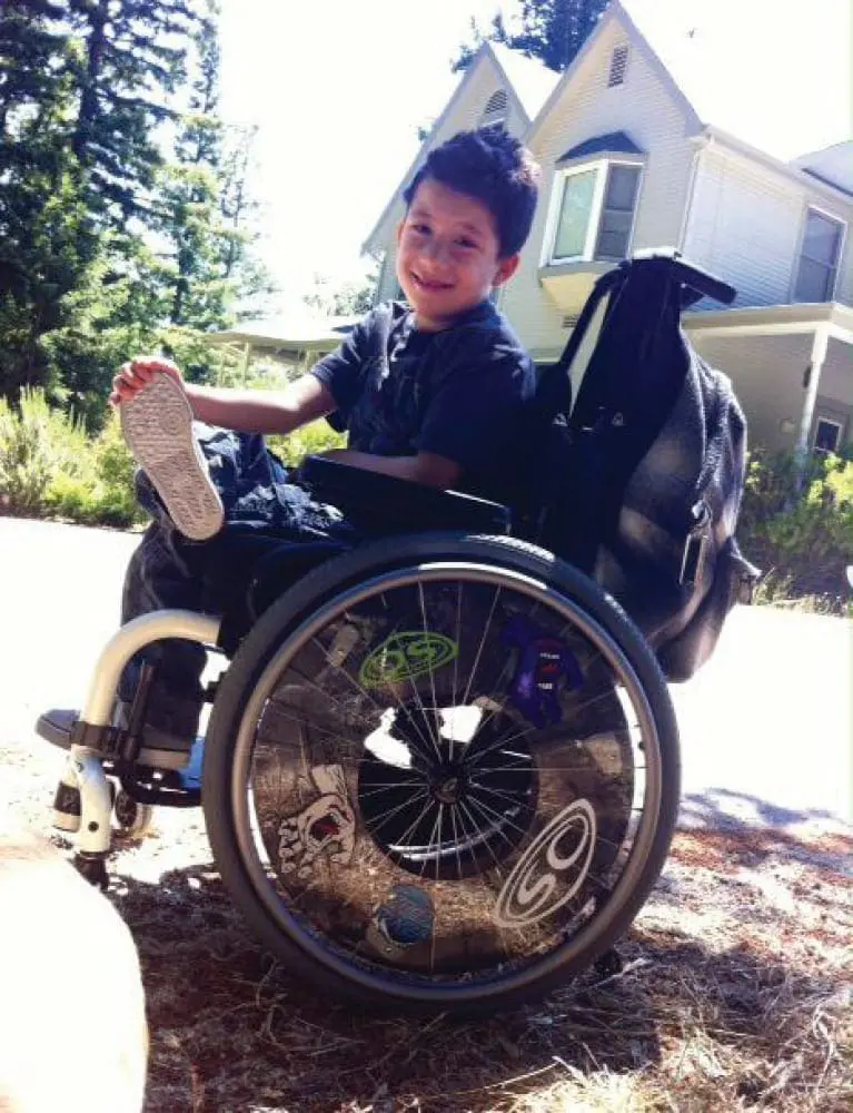 Joshy in his wheelchair with a house and trees in the background.