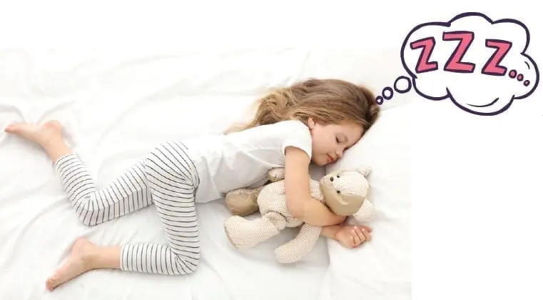 A young child sleeps soundly with a teddy bear on a white blanket.