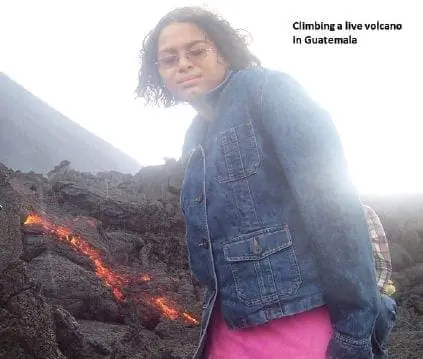 A woman wearing a denim jacket and pink skirt stands near a lava flow on a volcano.