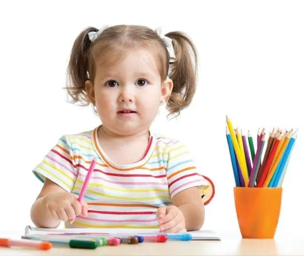 Toddler with pigtails holds a pink crayon at a table with colored pencils.