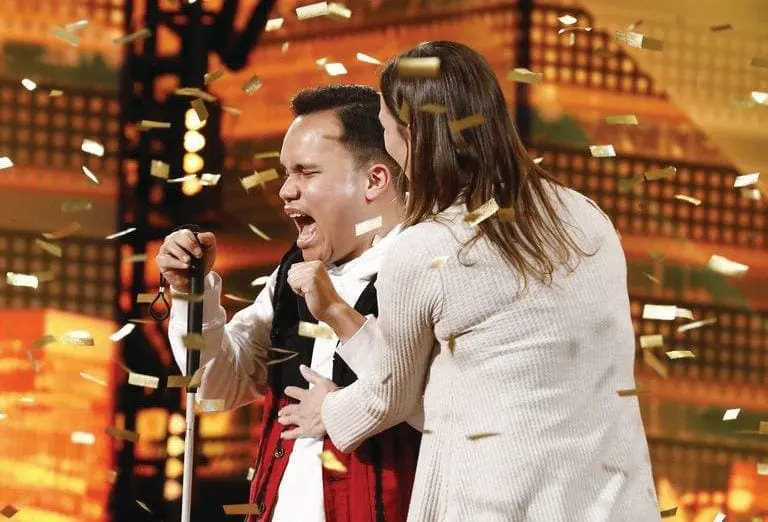 A man sings into a microphone with a woman embracing him as confetti falls.