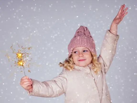 A young girl in a pink hat and coat holds a sparkler with one arm raised.