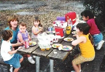A group of people, including a woman and several children, sit around a picnic table.