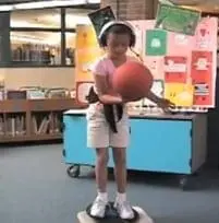 Young girl with headphones on, holding a ball, and standing on a balance board.