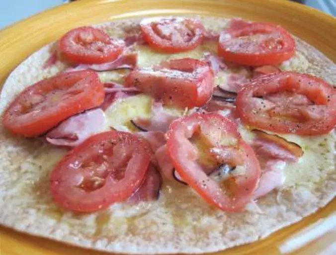 A round tortilla pizza is topped with sliced tomatoes, cheese, and ham.