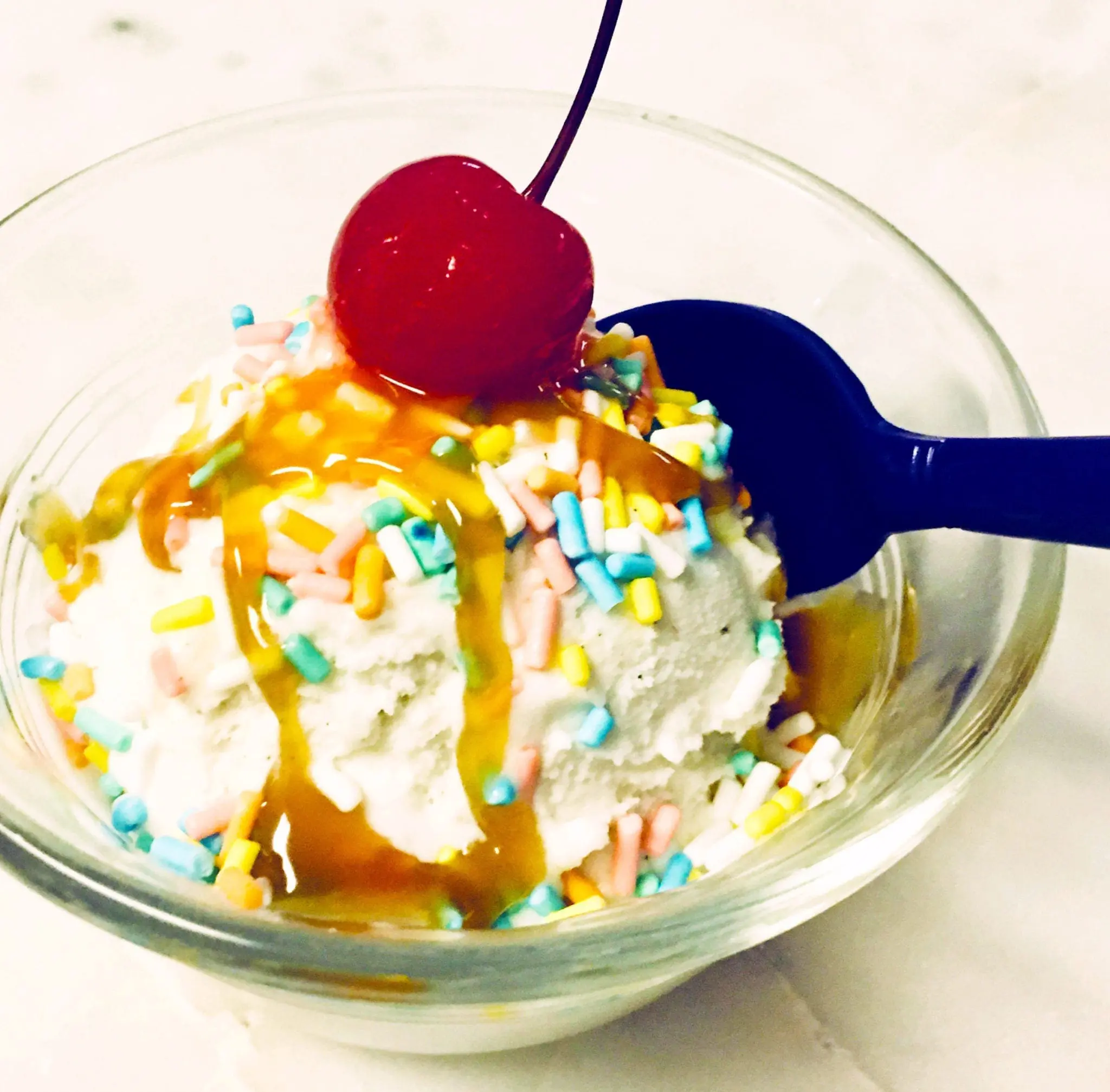 Scoop of ice cream in a glass bowl topped with sprinkles, caramel, and a cherry, with a spoon.