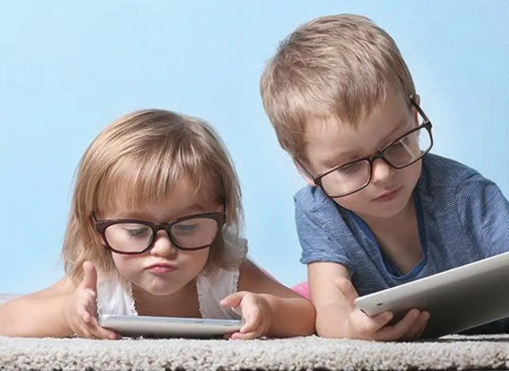 Two children wearing glasses look at separate tablets.