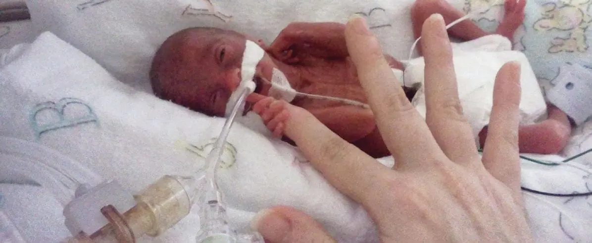 A preemie baby lies in an incubator with medical tubes while a hand reaches toward it.