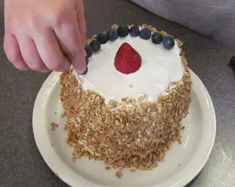 A hand places blueberries on top of a frosted watermelon cake decorated with granola.