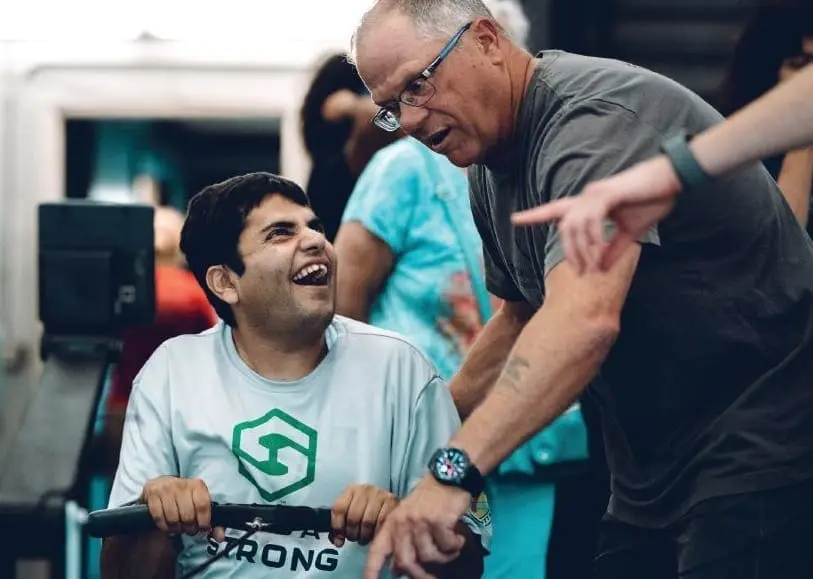 A man in a wheelchair laughs while looking at an older man assisting him with a workout.