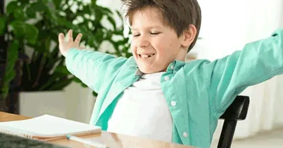 A young boy with his arms outstretched sits at a desk with a book open in front of him.