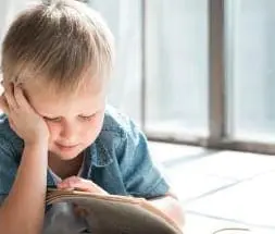 A young boy with blonde hair rests his head on his hand while reading a book.