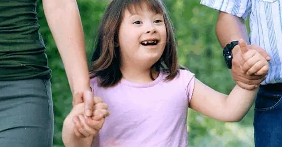A young girl with Down syndrome smiles while holding hands with two adults.