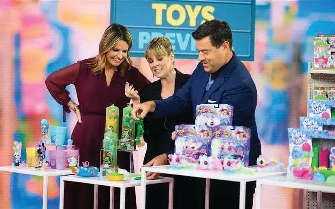 Three people stand behind a table displaying toys with a "TOYS PREVIEW" sign in the background.