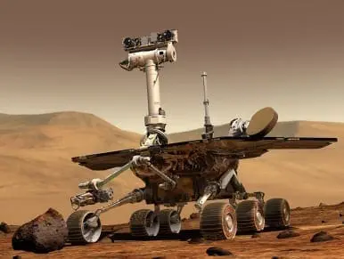 A Mars rover with six wheels sits on a dusty, rocky planet surface under a hazy sky.