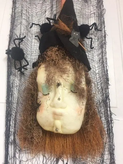 A Halloween decoration featuring a doll's face wearing a witch hat and surrounded by black netting and spiders.