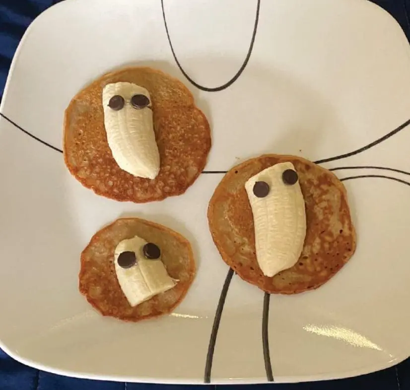 Three pancakes decorated to look like ghosts using banana slices and chocolate chips.