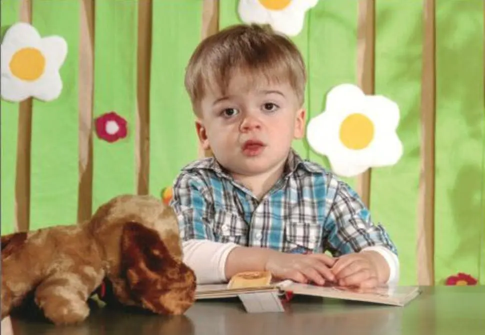 Young boy with a stuffed dog sits at a table in front of a colorful background.