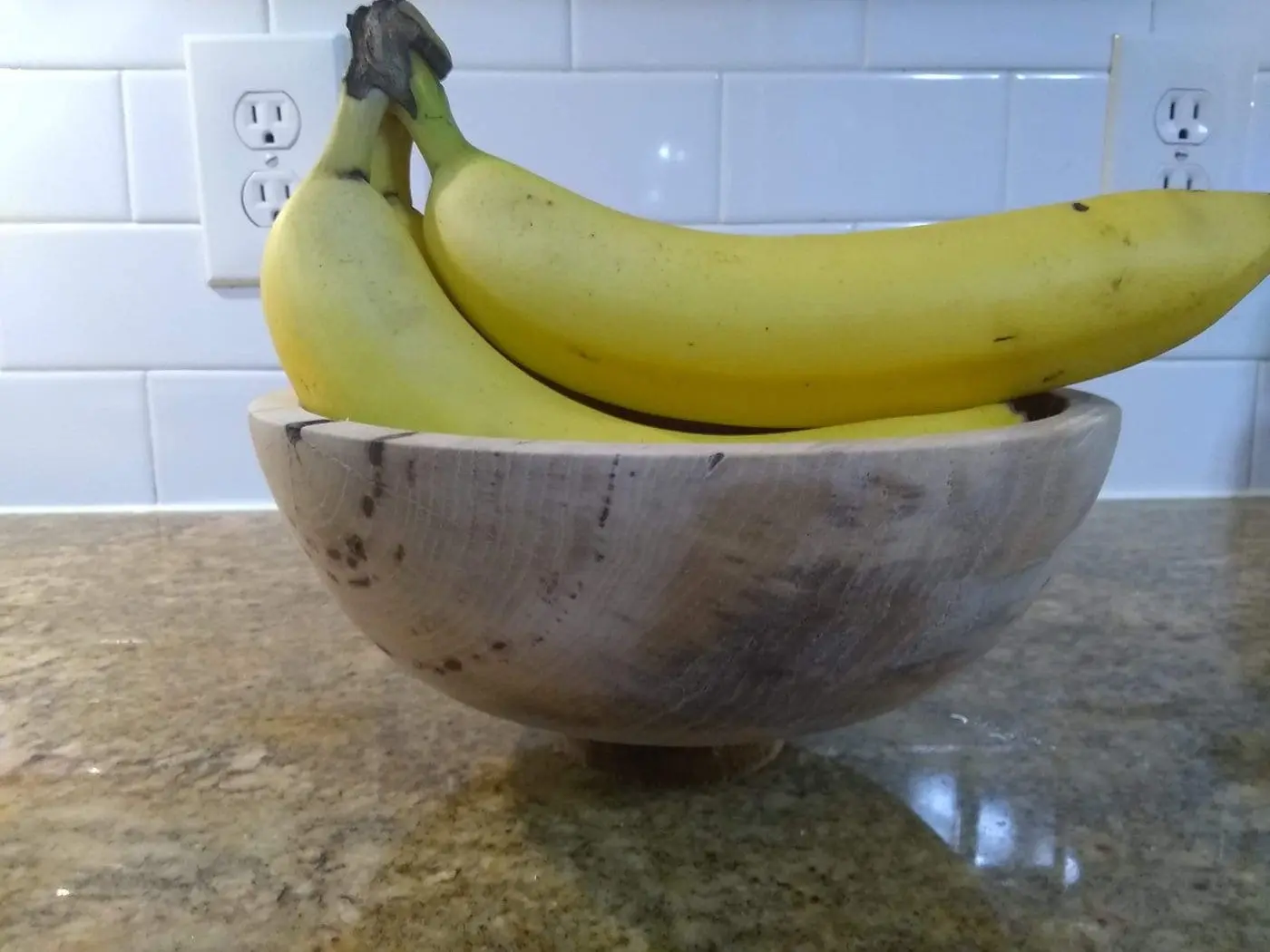 Three ripe bananas in a wooden bowl on a speckled countertop.