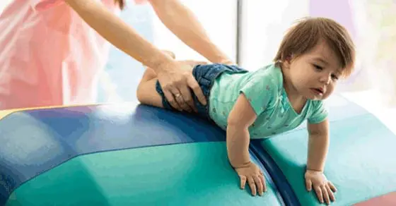 A child crawls on a soft, colorful play structure with an adult's hands supporting them.