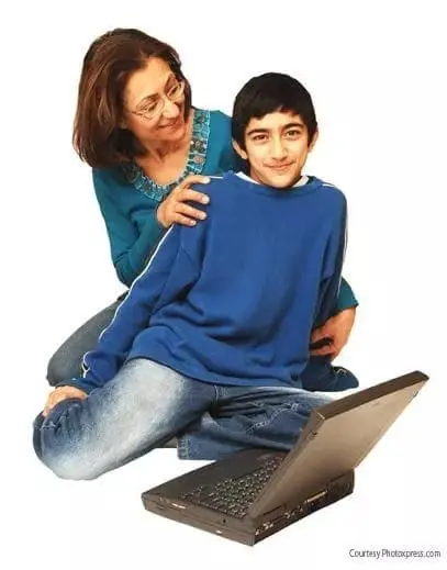 Woman with glasses and a blue shirt helps a young boy in a blue shirt with a laptop.