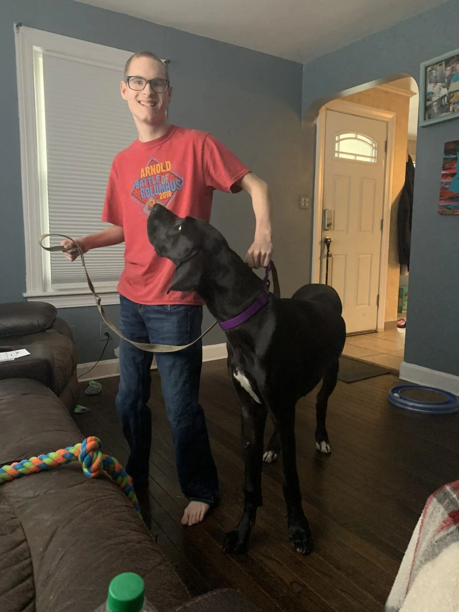A person in a red shirt and jeans stands with a large black dog on a leash.