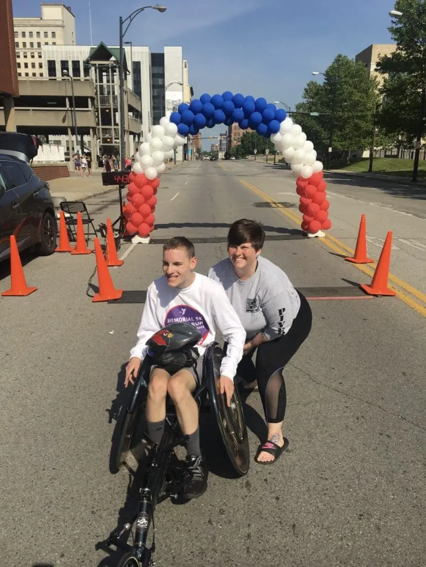 A boy in a wheelchair and a woman stand under a balloon arch at the start of a race.