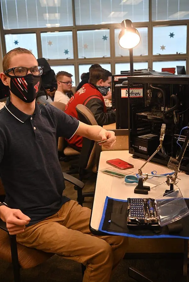 A man wearing glasses and a face mask gestures while sitting at a computer.