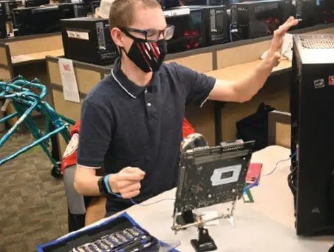Cody wearing a mask and glasses, gesturing toward a computer while working at a desk.