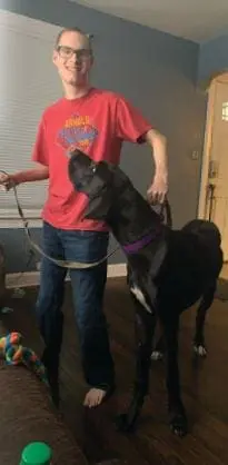 Cody, wearing glasses and a red shirt, holds a leash attached to a black dog.