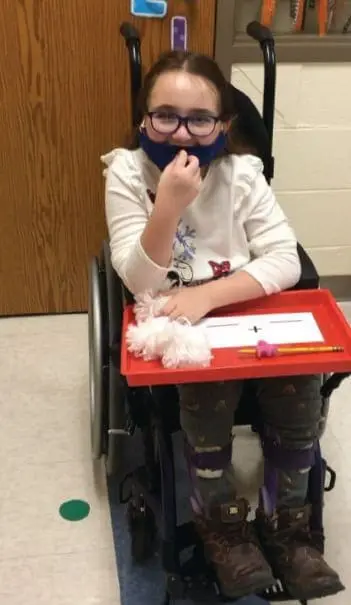 A young person in a wheelchair wears a mask and glasses, with craft supplies on a tray in front of them.