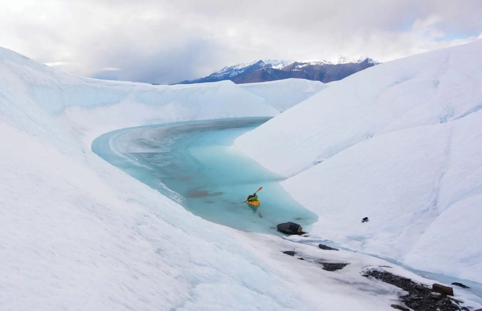 Person in a yellow kayak navigates a bright blue glacial pool surrounded by snow-covered mountains.
