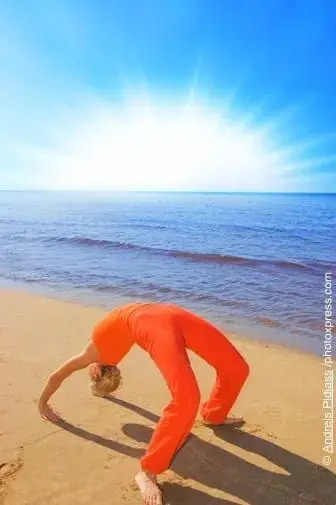 Person in orange yoga pants doing a backbend on a sandy beach with the ocean and bright sun in the background.