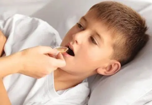 A person gives a young boy medicine on a spoon while he lies in bed.