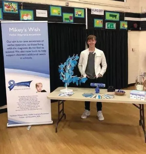 Mikey stands by an awareness banner at a quiz night with a table of displays.