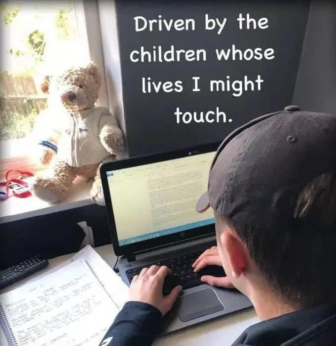 A young person wearing a baseball cap works at a laptop, with a teddy bear visible through a window in the background.