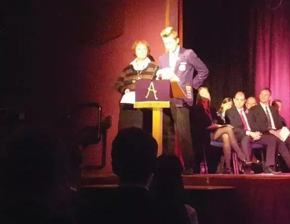 Mikey, a young man with blond hair, stands on a stage at a podium with an adult, reading a poem.