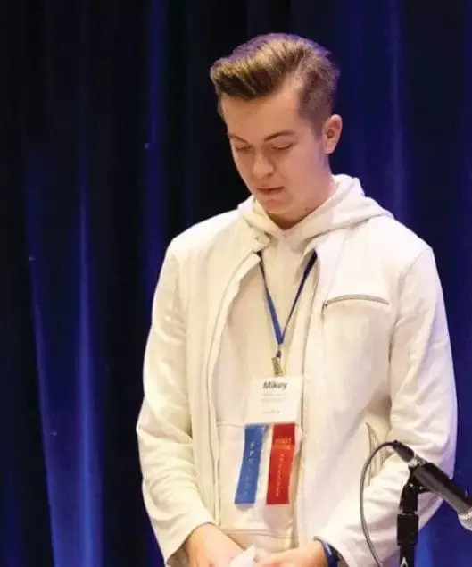Mikey looking down while wearing a white jacket and lanyard with a name tag.