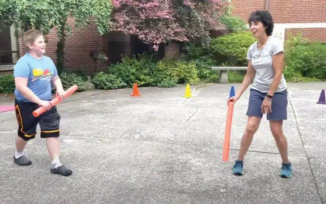 Two people hold pool noodles while standing in a paved outdoor area with cones.