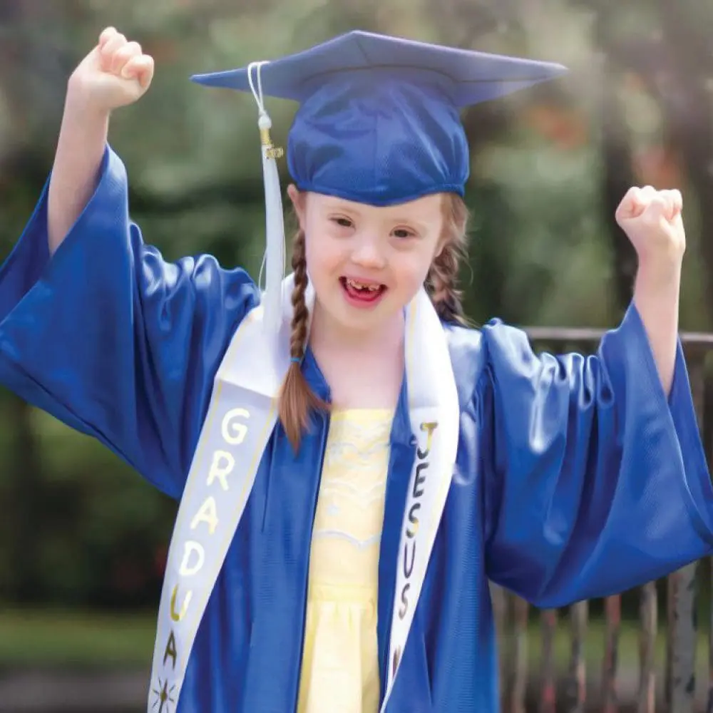 A young graduate in a blue cap and gown with arms raised in celebration.
