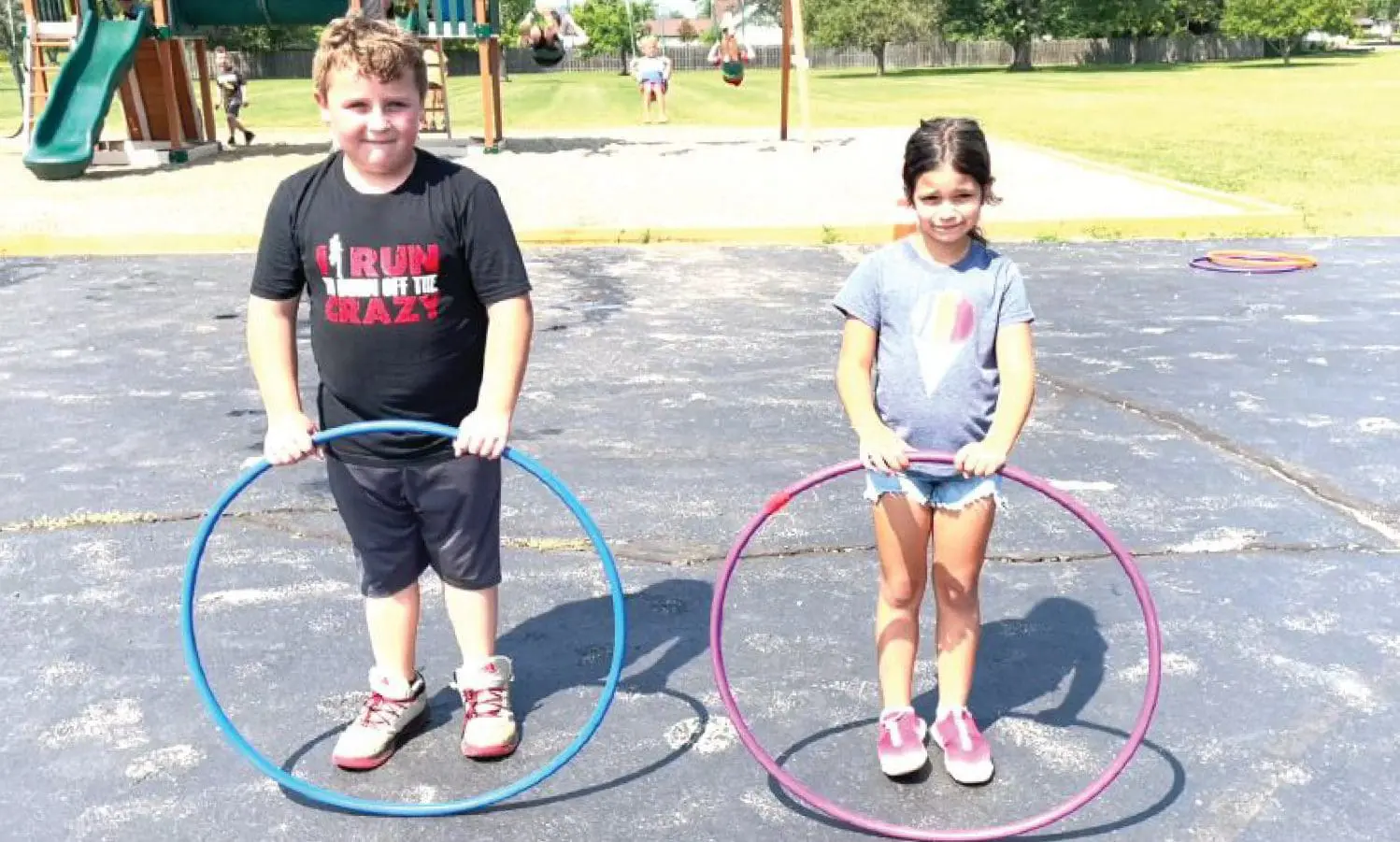 Two children stand holding hula hoops on a paved surface with playground equipment in the background.