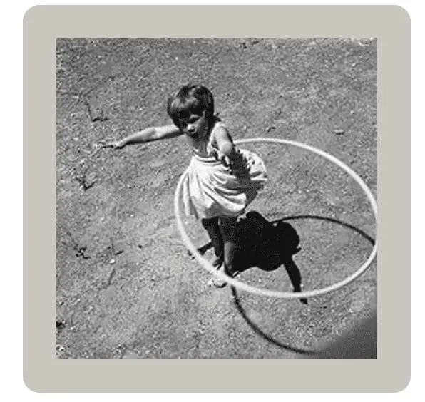 Young child in a dress twirling a hula hoop on a paved surface.