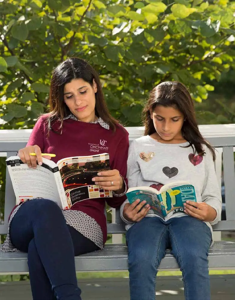 Two women sitting on a bench outdoors reading books.